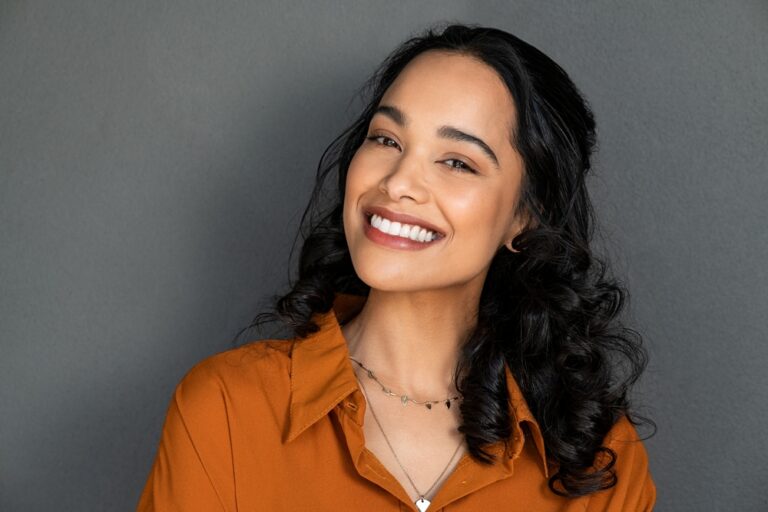 Smiling young woman with long curly dark hair wearing an orange shirt and layered necklaces, standing in front of a plain gray background.